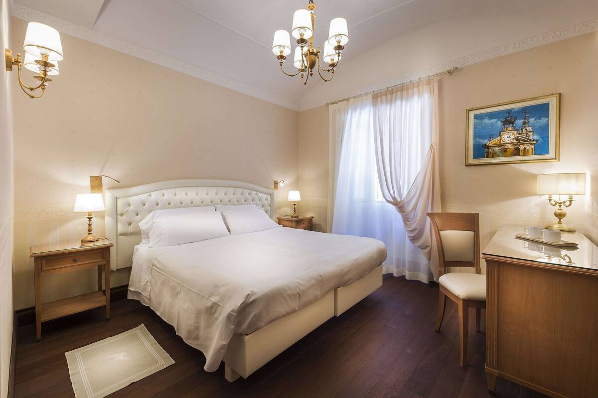 A hotel room with a large bed, bedside lamps, a desk with a lamp, a chair, and a painting on the wall.