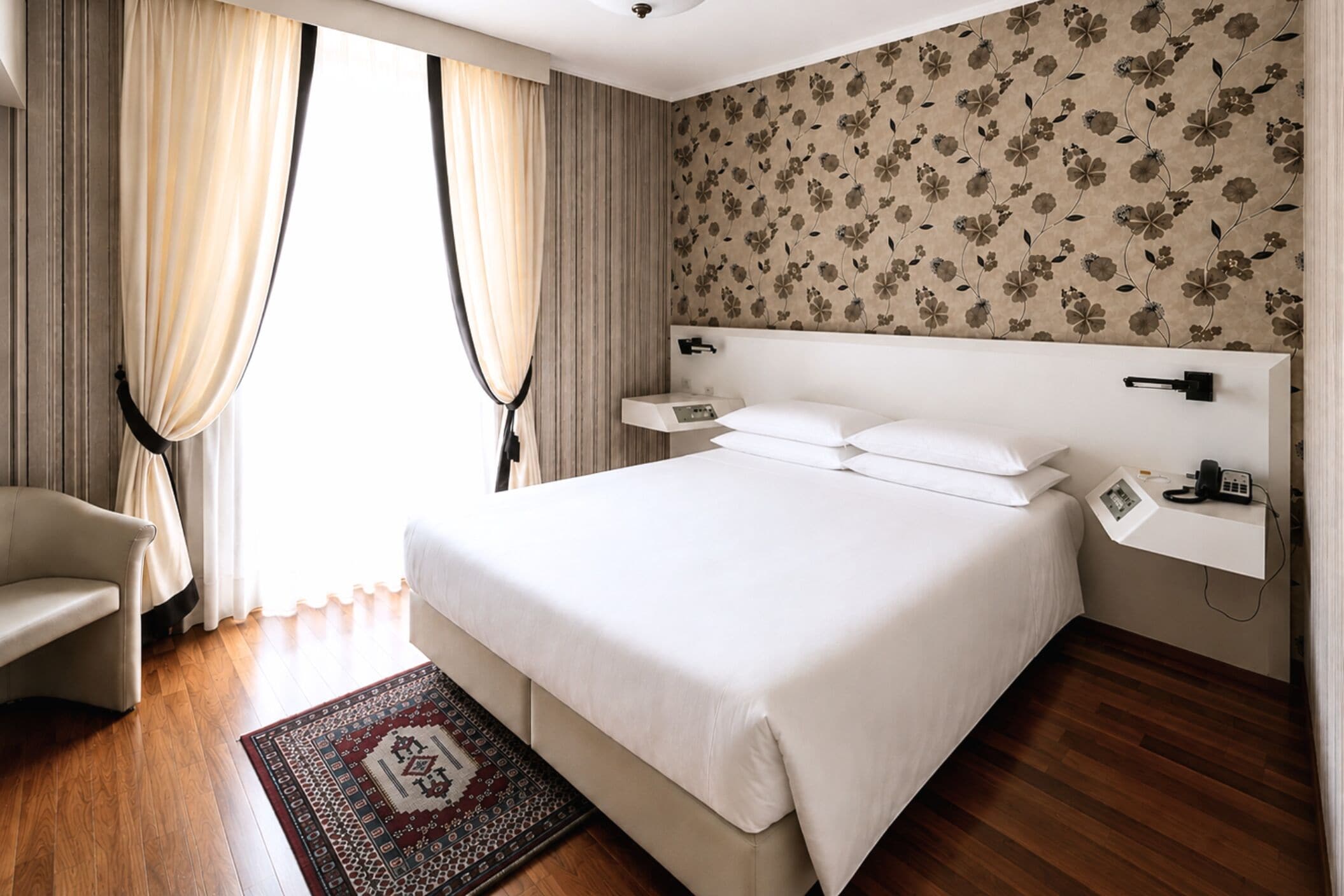 Habitación de hotel con una cama grande, mesitas de noche, una silla y un papel tapiz estampado.