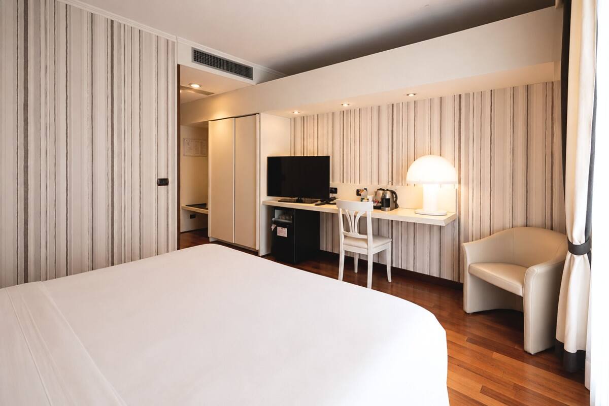 A hotel room with a large bed, a desk with a TV, a chair, and a lamp.