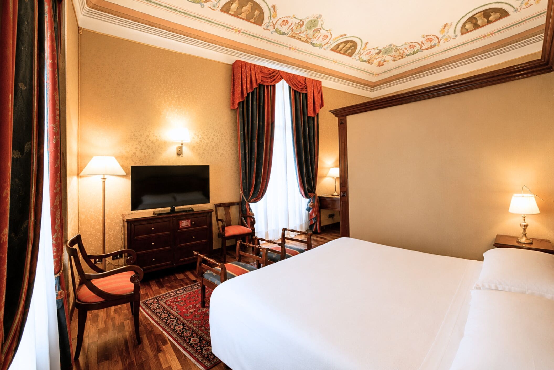 A hotel room with a bed, a television, a desk with a lamp, a chair, and a window with curtains.