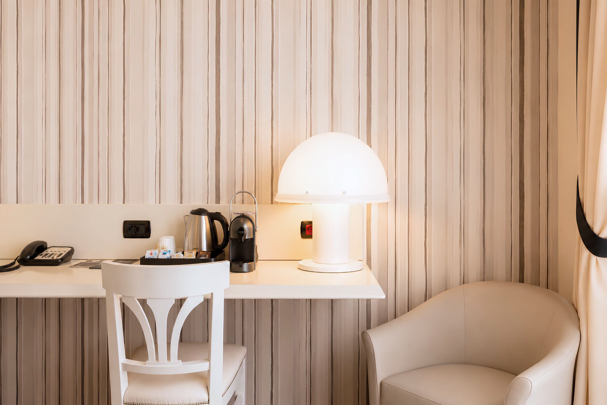 Ett hotellrum med randig tapet, ett skrivbord med en lampa, en stol, en telefon och en kaffebryggare.