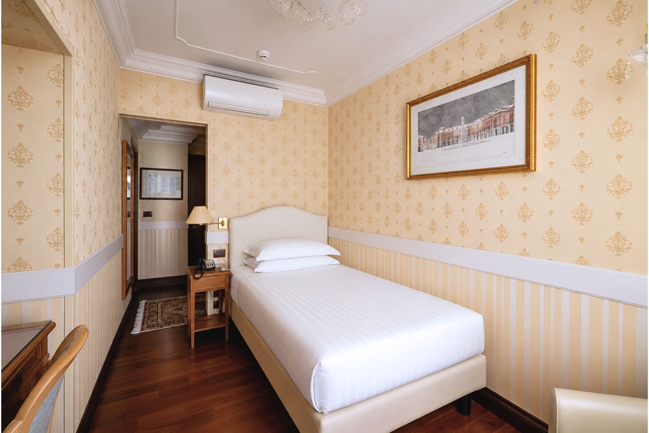 A hotel room with a bed, a nightstand, a lamp, a framed picture on the wall, and a door leading to another room.
