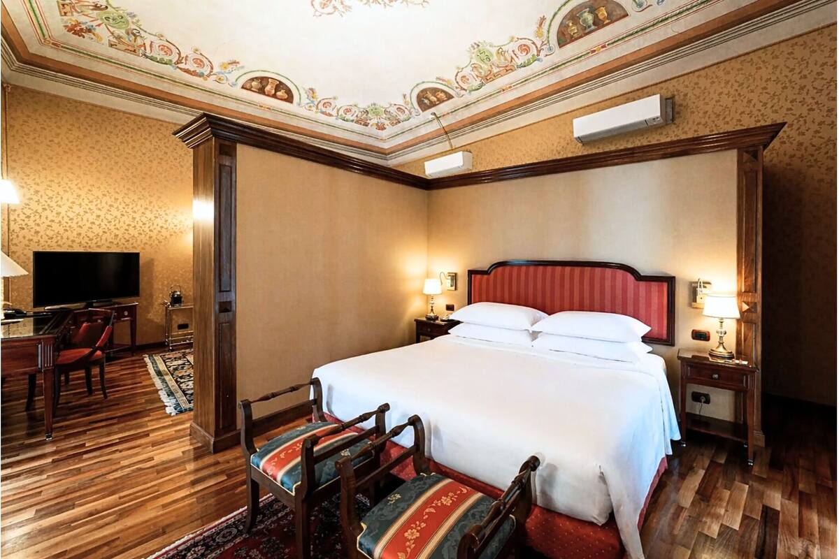 A hotel room with a large bed, two bedside tables with lamps, a TV, a desk with a chair, and a patterned rug.