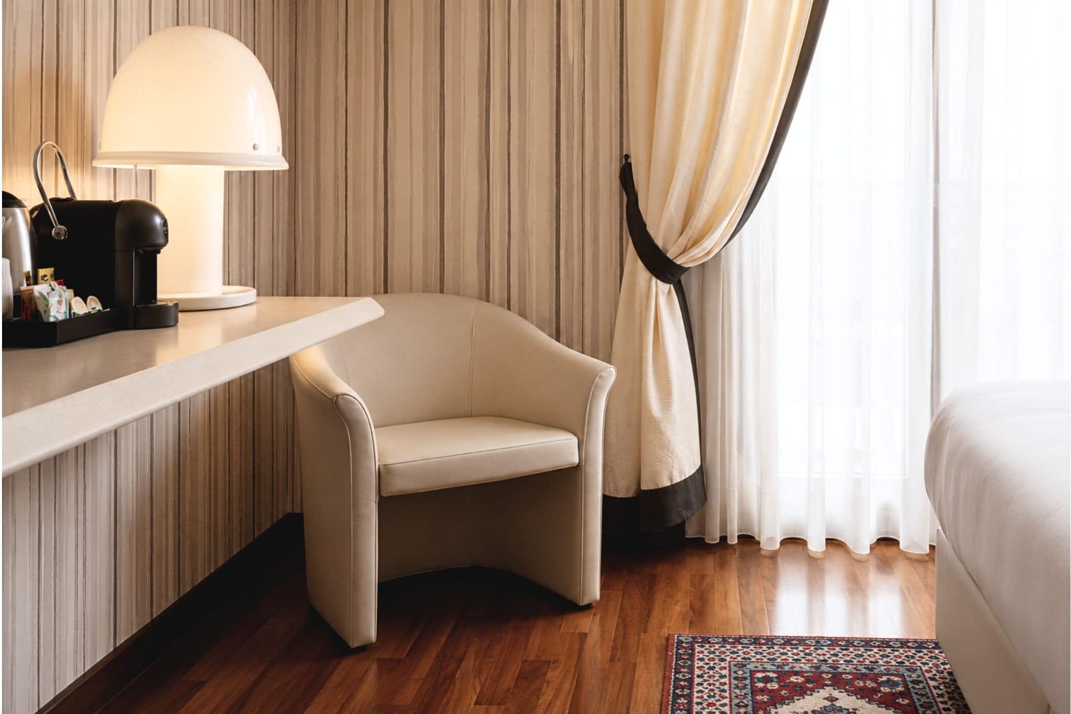 Una habitación de hotel con sofá, escritorio con lámpara, cafetera y alfombra estampada.