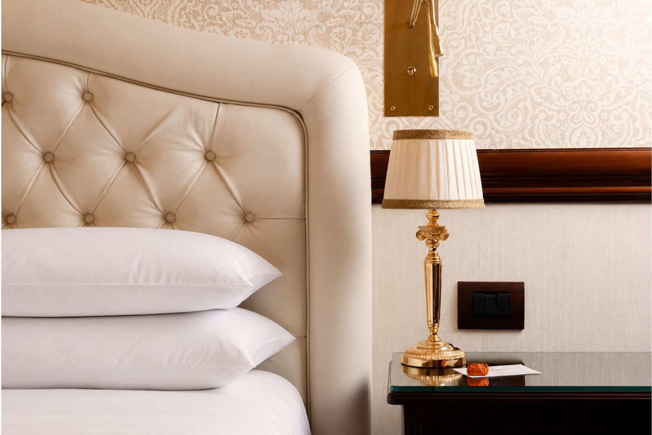 A hotel room with a tufted headboard, a bedside lamp, a nightstand, and a wall switch.