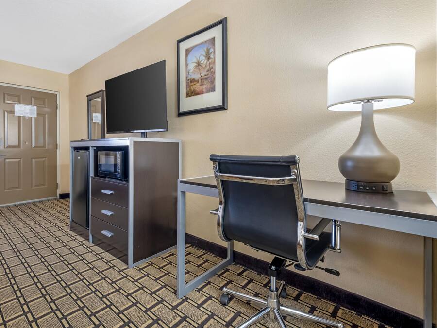 A hotel room with a desk, chair, television, minibar, and a patterned carpet.
