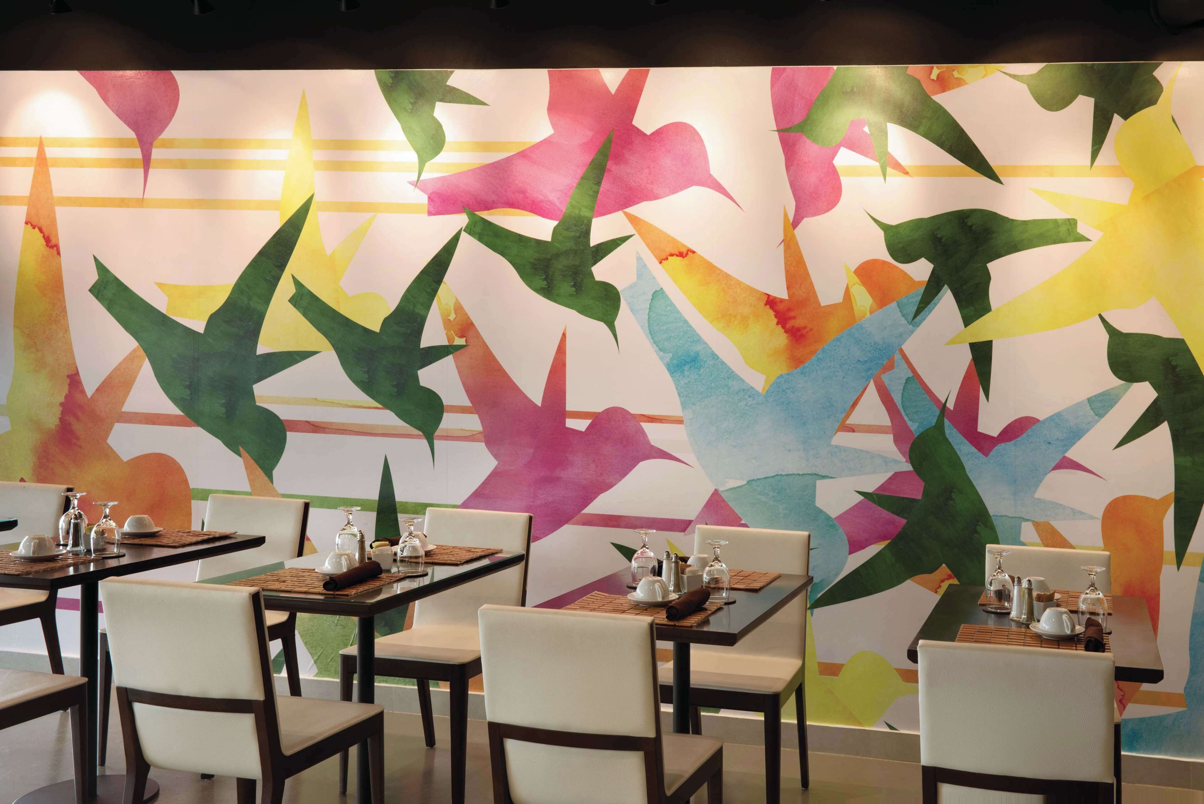 Un restaurant dont les tables sont dressées pour le service, avec une fresque murale abstraite et colorée.