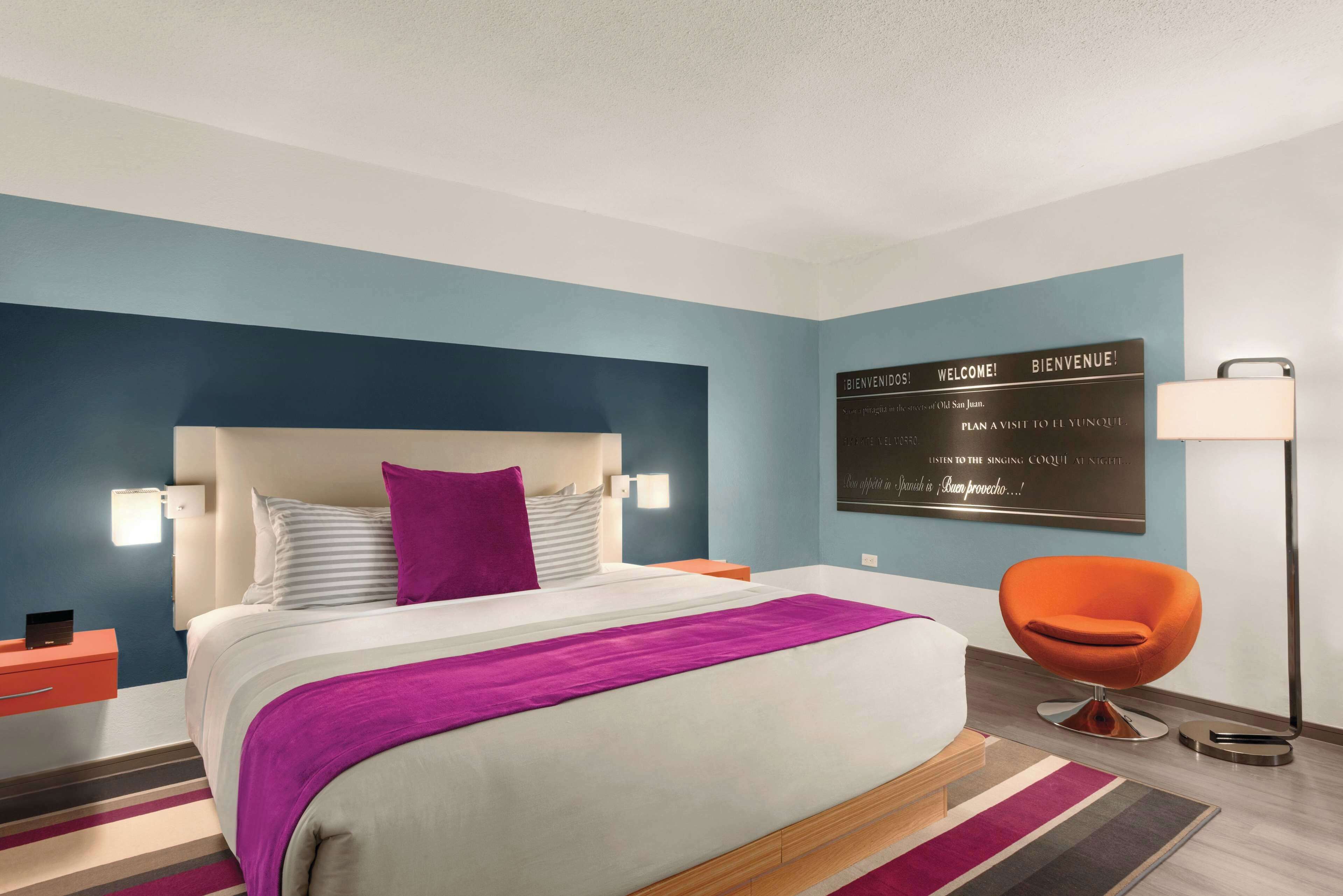 A modern hotel room with a large bed, a nightstand, a chair, and a wall-mounted sign.