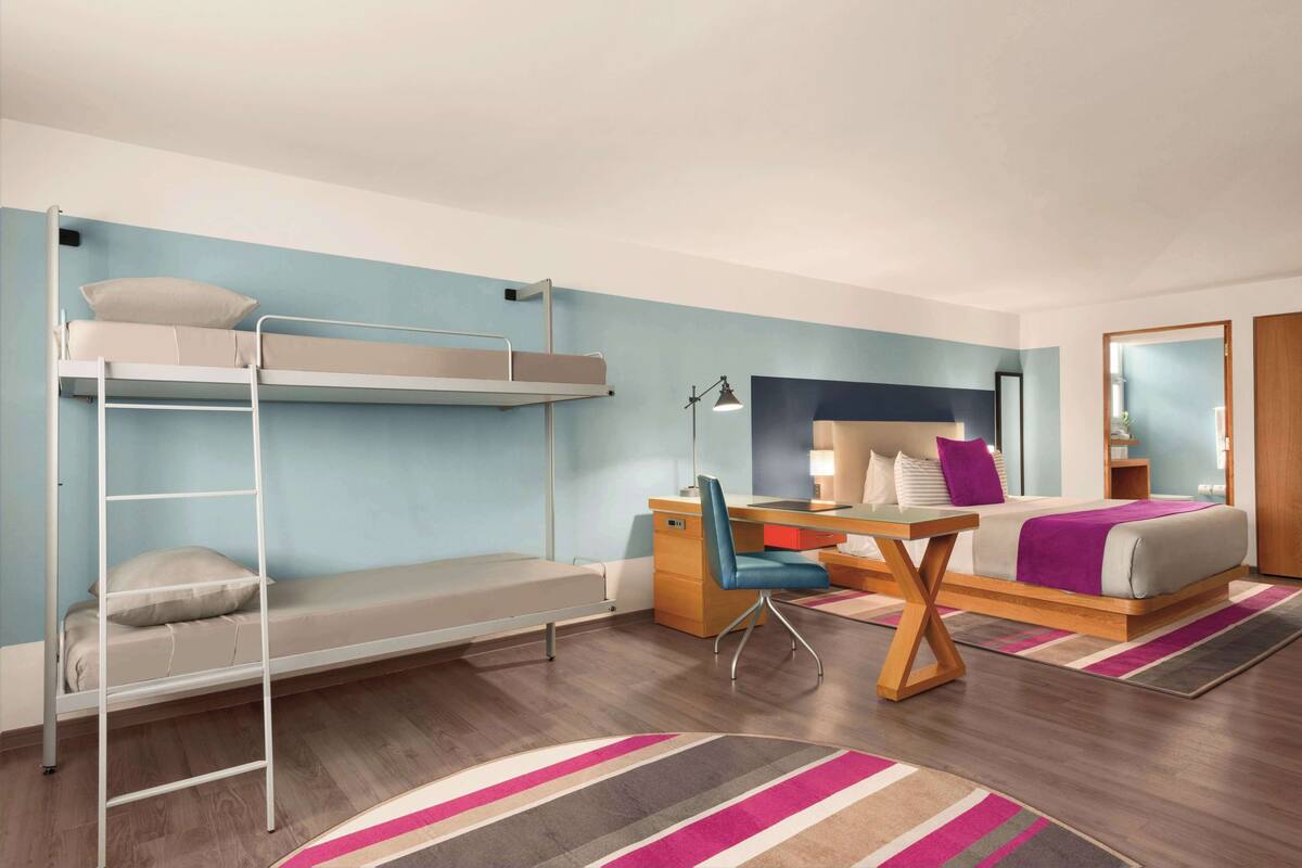 A hotel room with a bunk bed, a desk with a chair, a bed with a purple blanket, and a mirror.