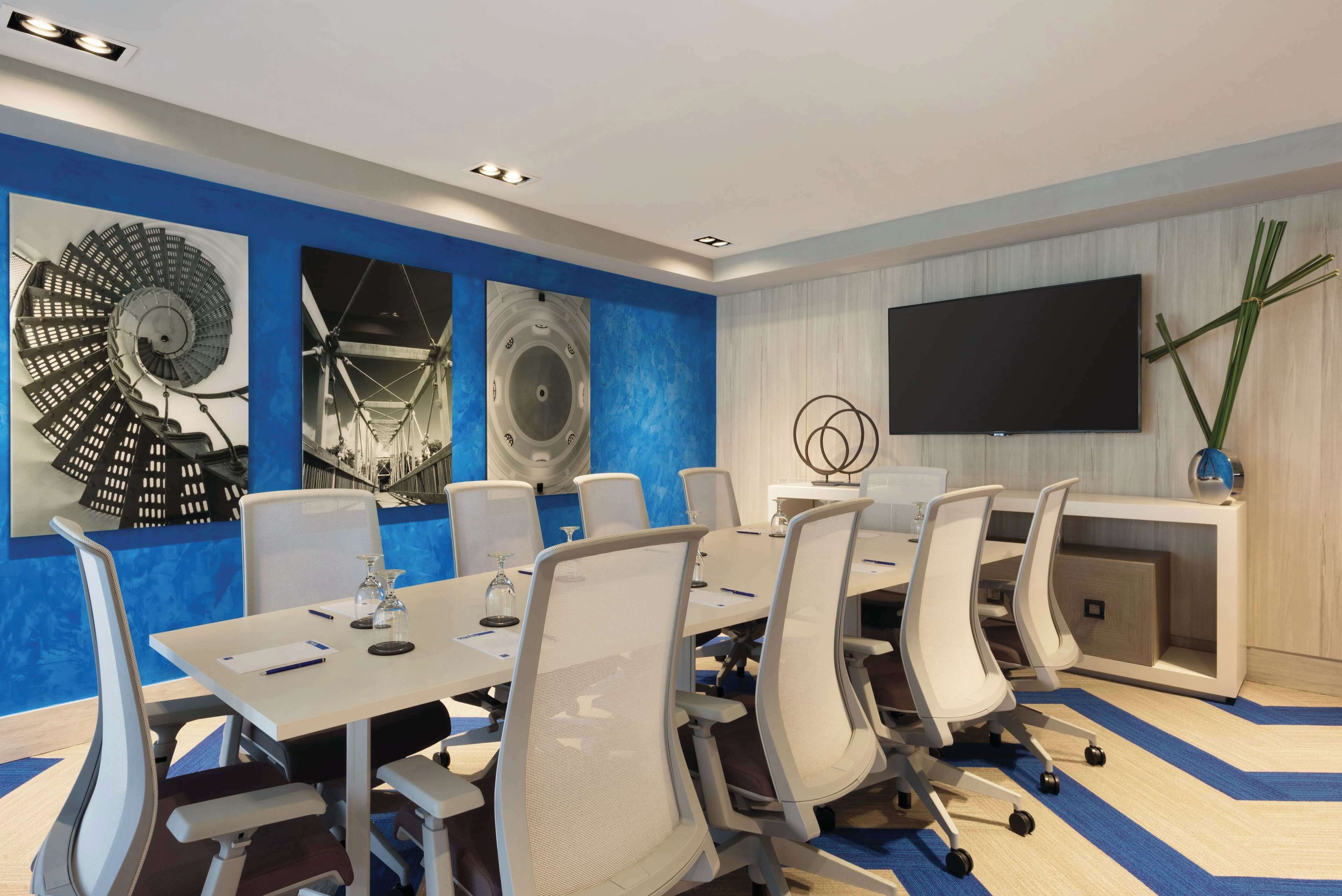 A modern conference room with a large table, white chairs, and a flat-screen TV on the wall.