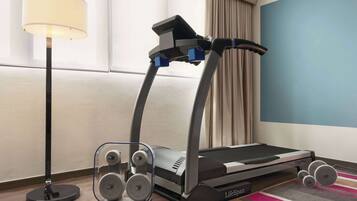 A treadmill in a hotel room with a lamp, a window with curtains, and a striped rug.