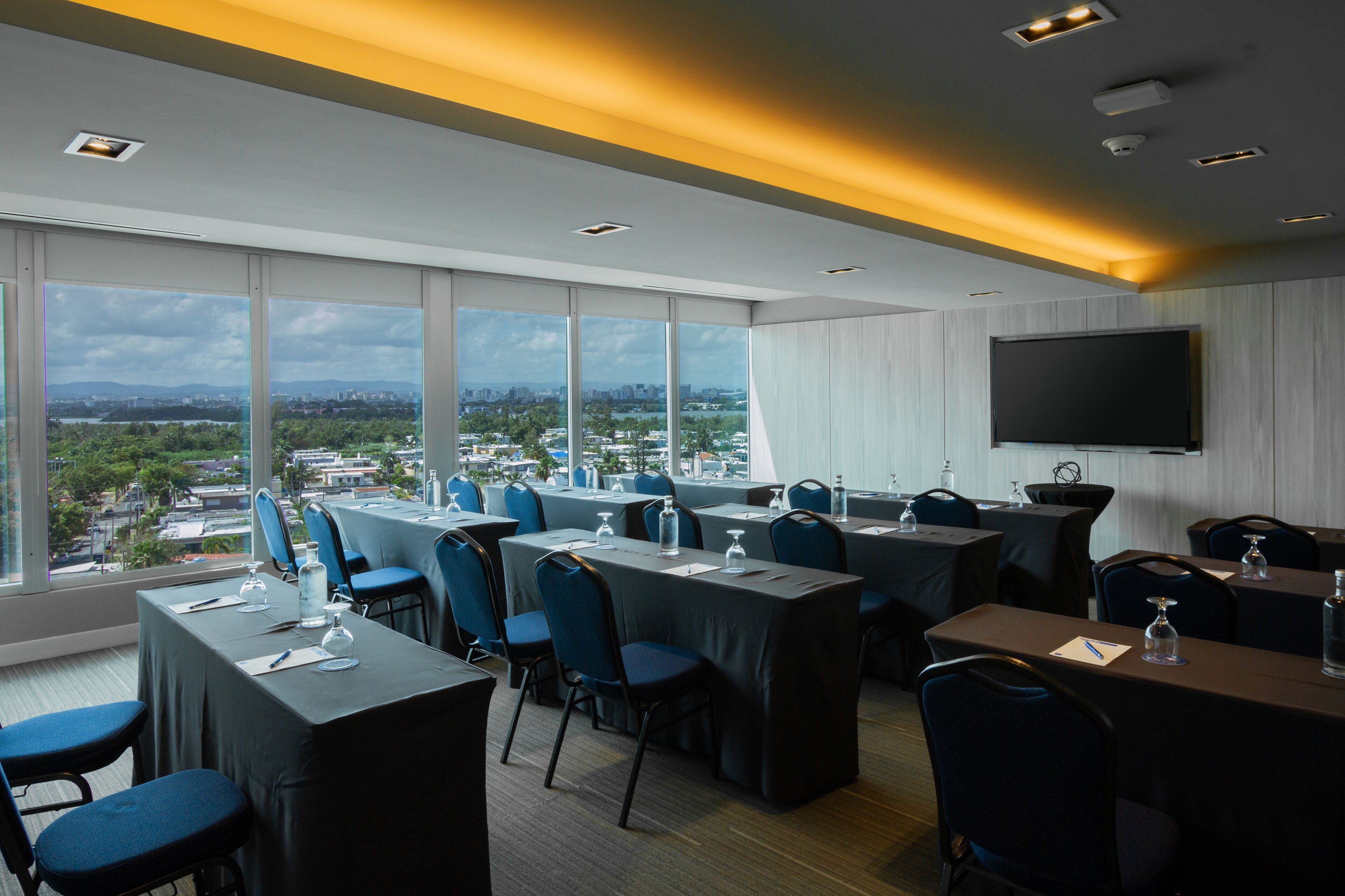 A conference room with large windows offering a city view, multiple tables with chairs, and a flat-screen TV on the wall.