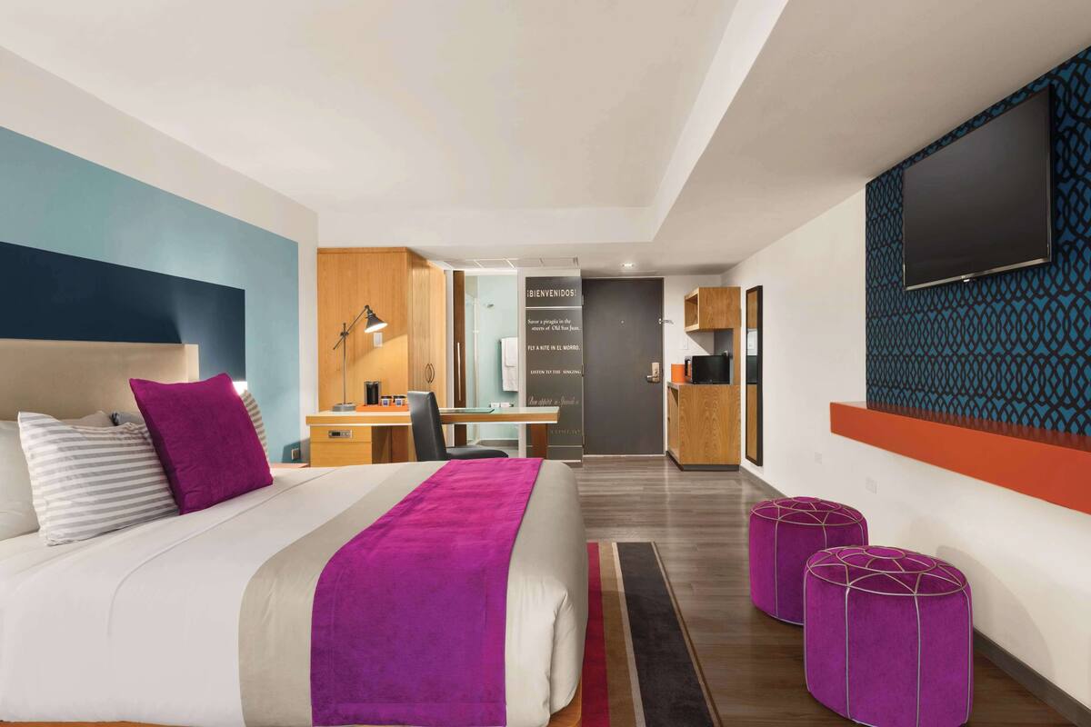A modern hotel room with a bed, a desk, a TV, and a kitchenette.