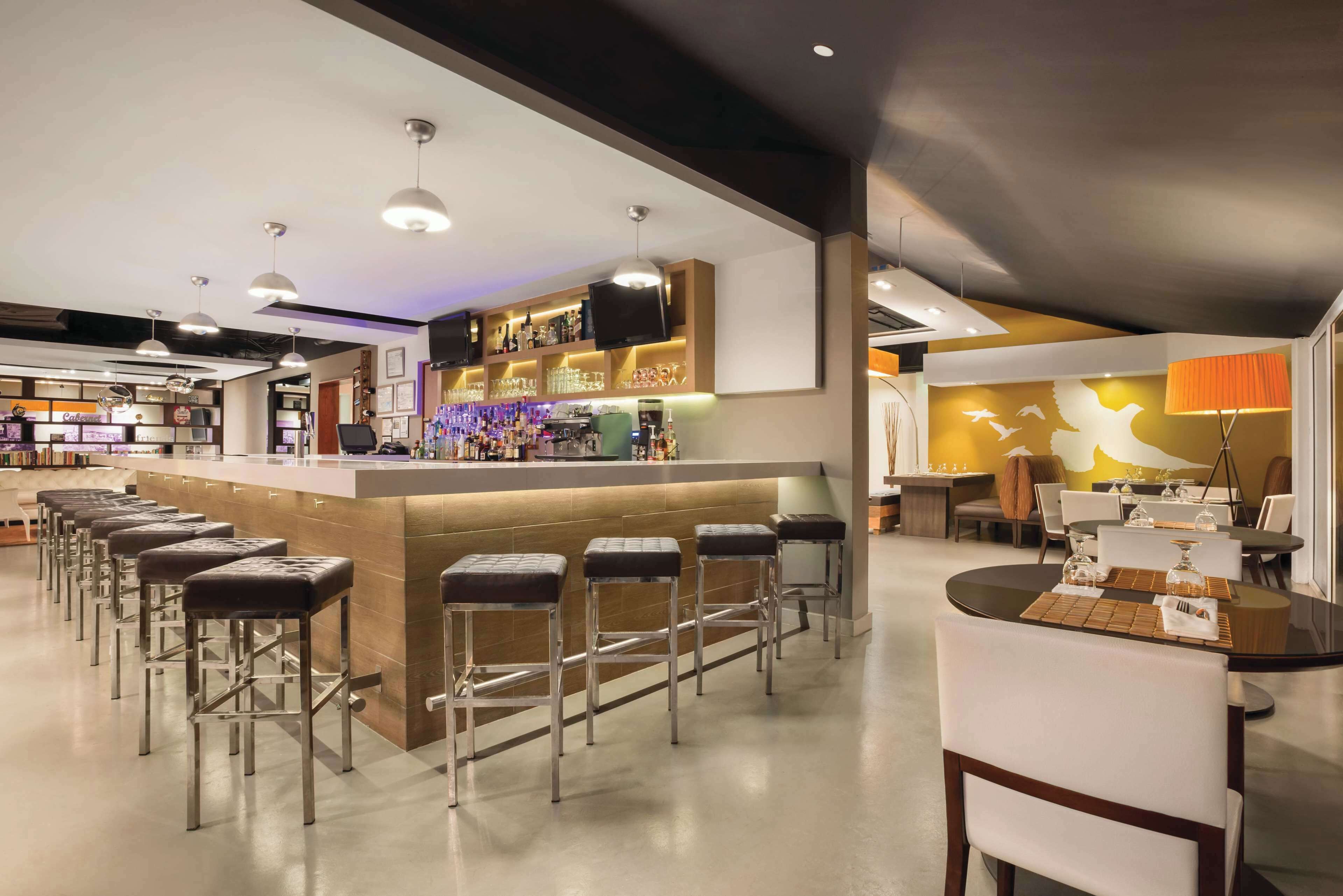 A modern bar area with a long counter, multiple stools, and overhead lighting.