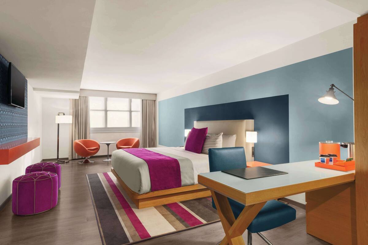 A modern hotel room with a large bed, a desk, and a chair. There is a TV mounted on the wall, a window with curtains, and a lamp on the desk.