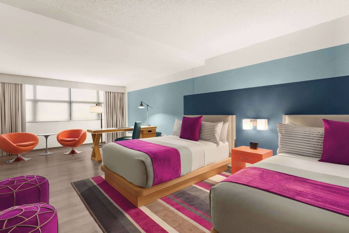 A modern hotel room with a large bed, two bedside tables, a desk, and two orange chairs.