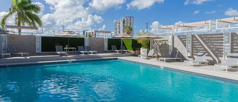 A swimming pool with lounge chairs and umbrellas, surrounded by a building with a palm tree and a clear sky.