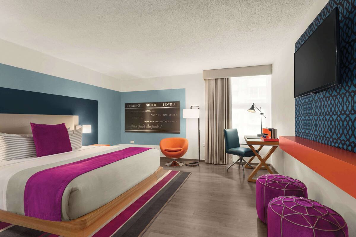 A modern hotel room with a large bed, a desk, a chair, and a TV mounted on the wall.