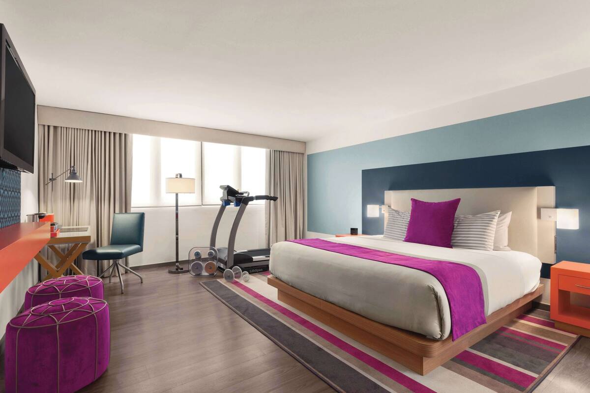 A hotel room with a bed, a desk, a treadmill, and ottomans.