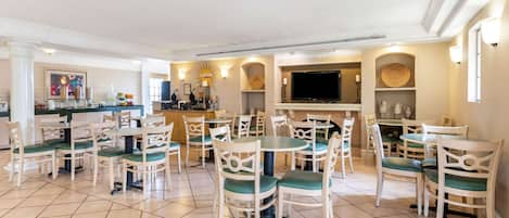 Un comedor amplio con sillas blancas y cojines verdes, suelo de baldosas y una zona de bar con televisor.