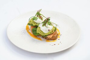 A white plate with a sandwich topped with avocado, greens, and a poached egg, garnished with microgreens.