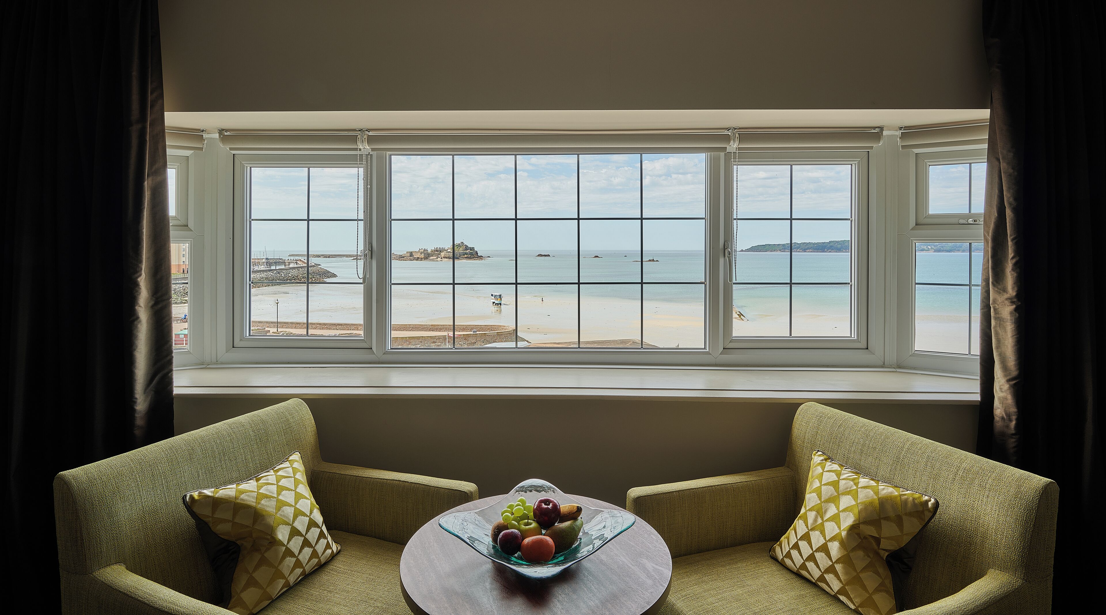 A room with a bay window overlooking a beach, two armchairs, and a table with fruit.