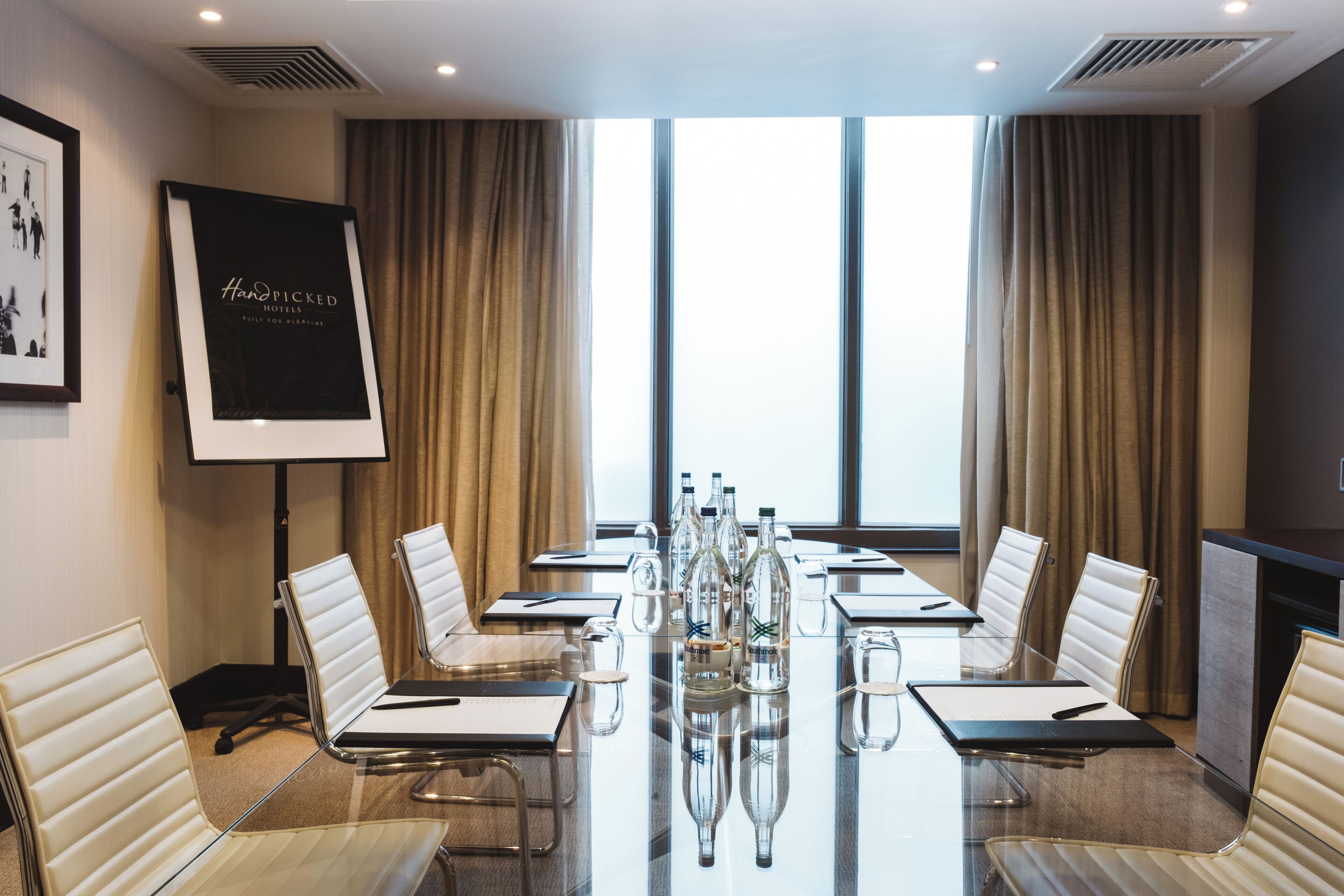 A conference room with a long table, white chairs, and a projector screen displaying 'Handpicked Hotels Built for Pleasure'.