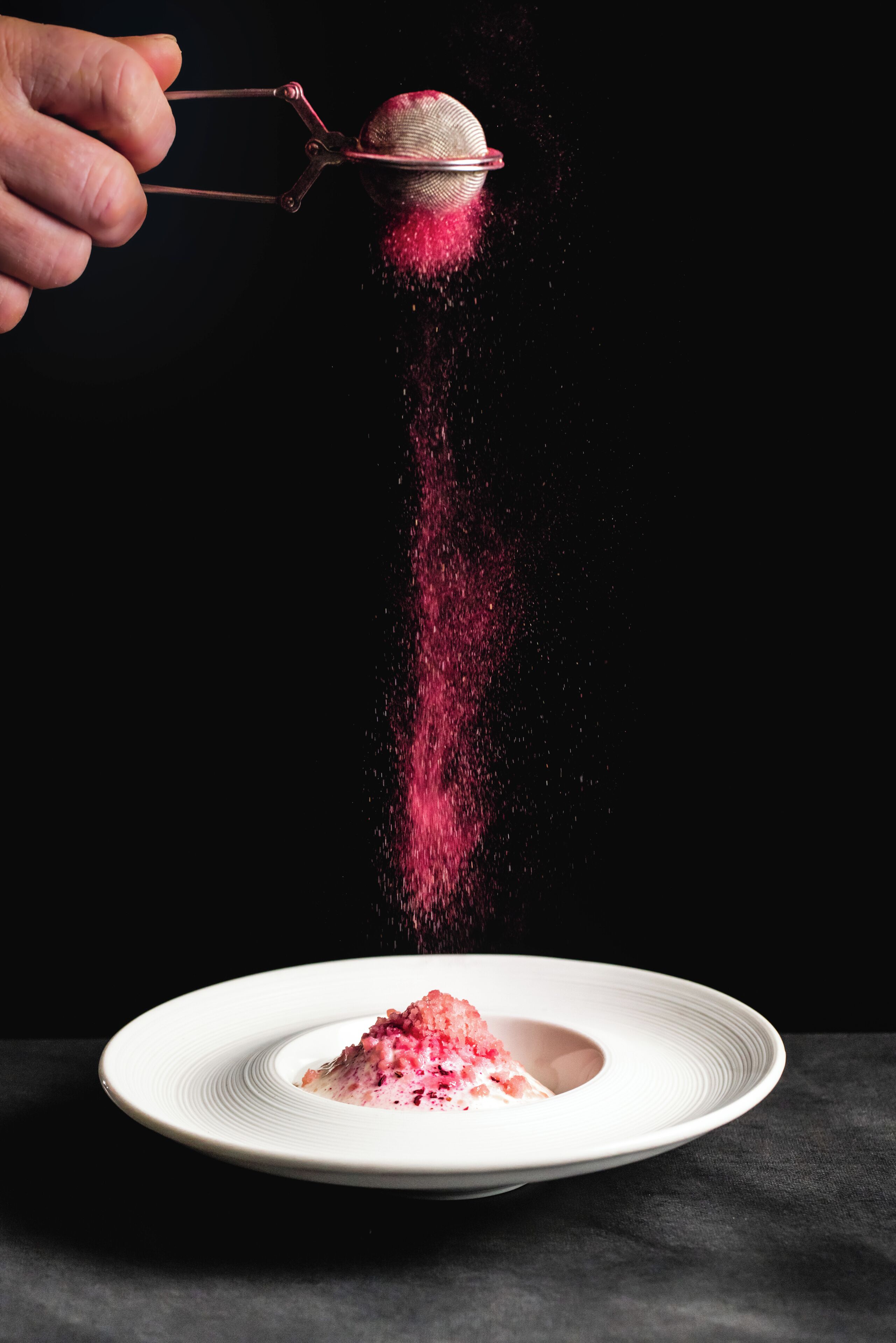 Une main tenant un tamis au-dessus d’une assiette contenant une substance poudreuse rose.