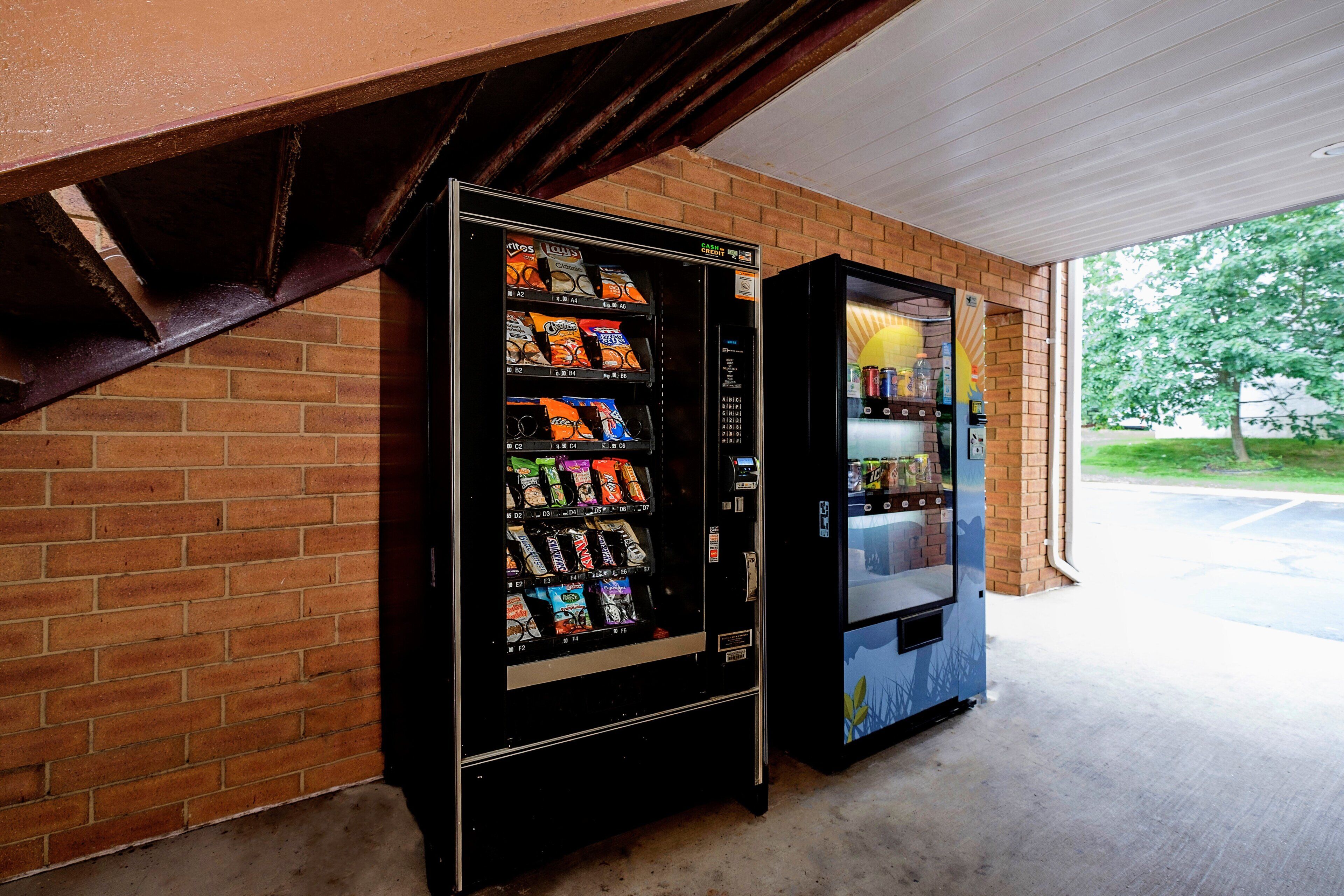 Two vending machines against a brick wall, one with snacks and the other with beverages.