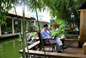 A man sitting on a wooden bench with a laptop, surrounded by a garden with a pond, bamboo, and various plants.