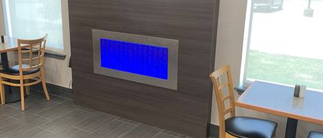 Un interior moderno con un televisor de pantalla plana montado en la pared, un panel azul iluminado debajo y una zona de comedor con sillas de madera y una mesa.