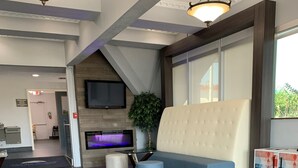A modern lobby with a grey tiled floor, a seating area with a sofa and ottoman, a flat-screen TV mounted on the wall, and a potted plant. - Howard Johnson by Wyndham Oklahoma City Airport/ Fairgrounds (Oklahoma City)