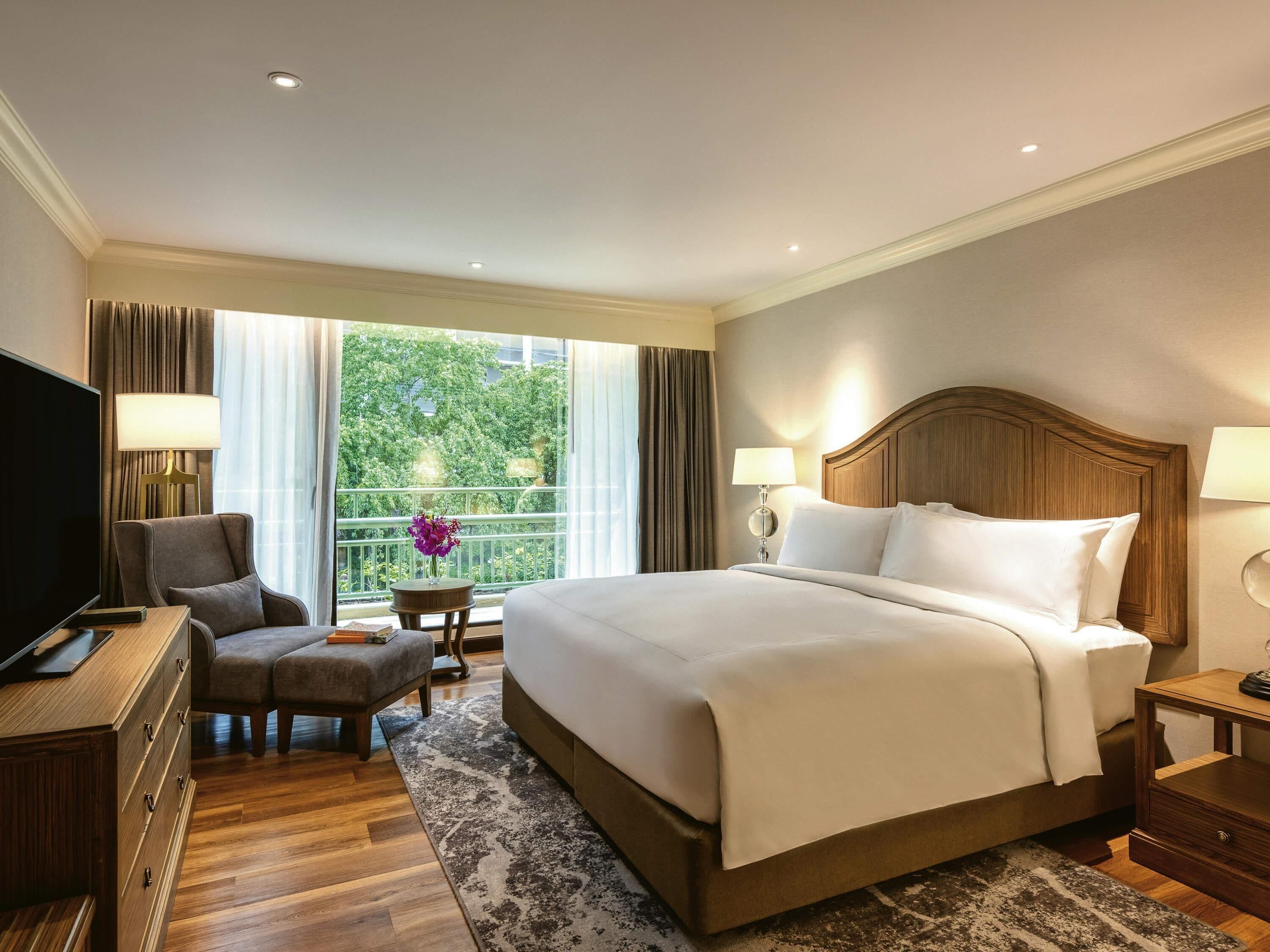 A hotel room with a bed, a TV, a desk, a chair, a small table with a vase, and a balcony with curtains.