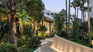 A curved pathway flanked by lush greenery and palm trees, leading to a modern building with large windows.