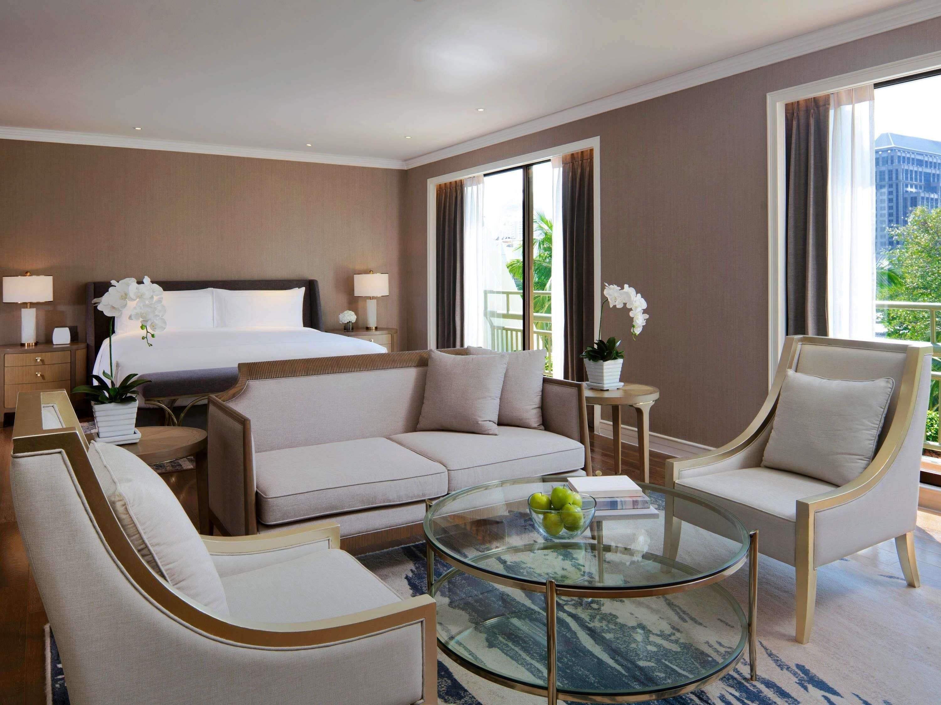 Ein Hotelzimmer mit einem Bett, einer Couch, zwei Sesseln, einem Glastisch mit Äpfeln und Blick auf Grünflächen.