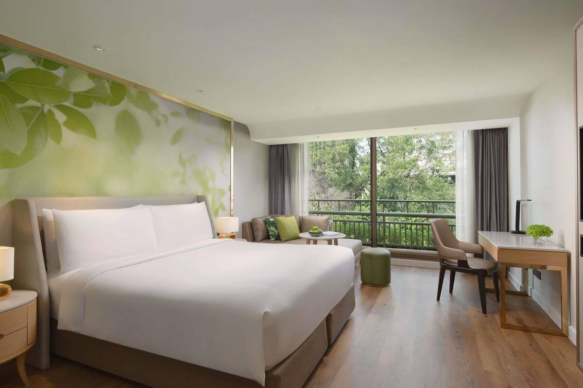 Ein modernes Hotelzimmer mit einem großen Bett, einem Schreibtisch, einem Sessel und einem Balkon mit Blick auf Grünflächen.