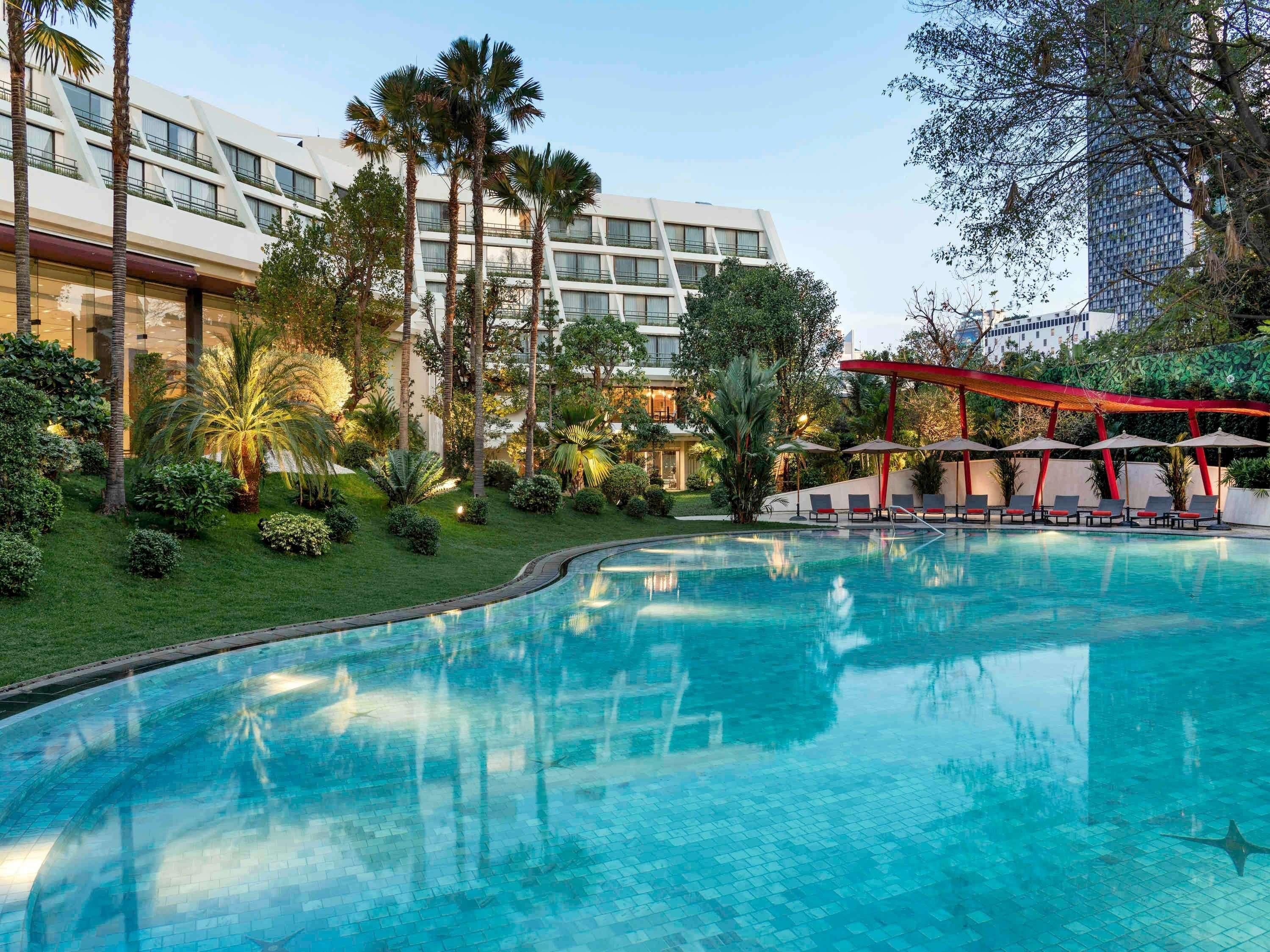 A swimming pool with lounge chairs and umbrellas, surrounded by greenery and modern buildings.