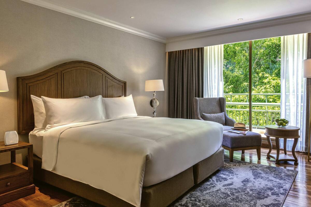 Ein Hotelzimmer mit einem großen Bett, einem Nachttisch, einem Sessel, einem kleinen Tisch und Blick auf Grün durch das Fenster.