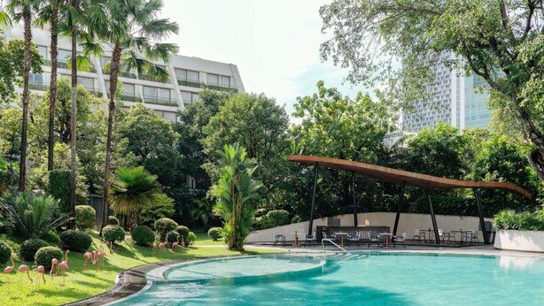 A swimming pool with a curved edge, surrounded by lush greenery and palm trees. There is a covered seating area with tables and chairs.