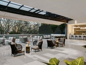 Un salon sur le toit avec des espaces de détente en plein air, un bar et une vue sur les arbres et les bâtiments.