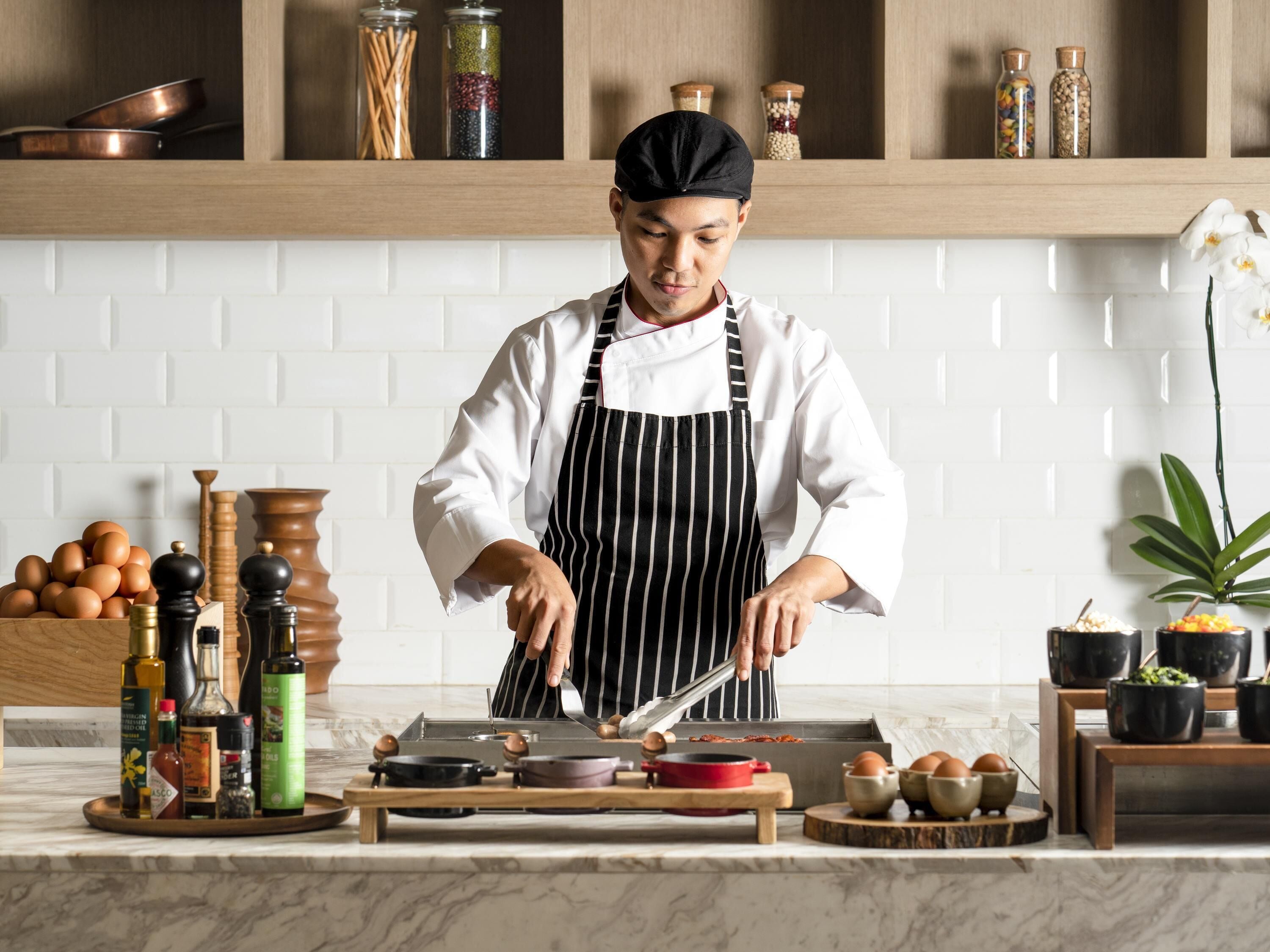 A chef in a kitchen with a white tiled backsplash, wooden shelves with jars, and a variety of cooking utensils and ingredients on the counter.