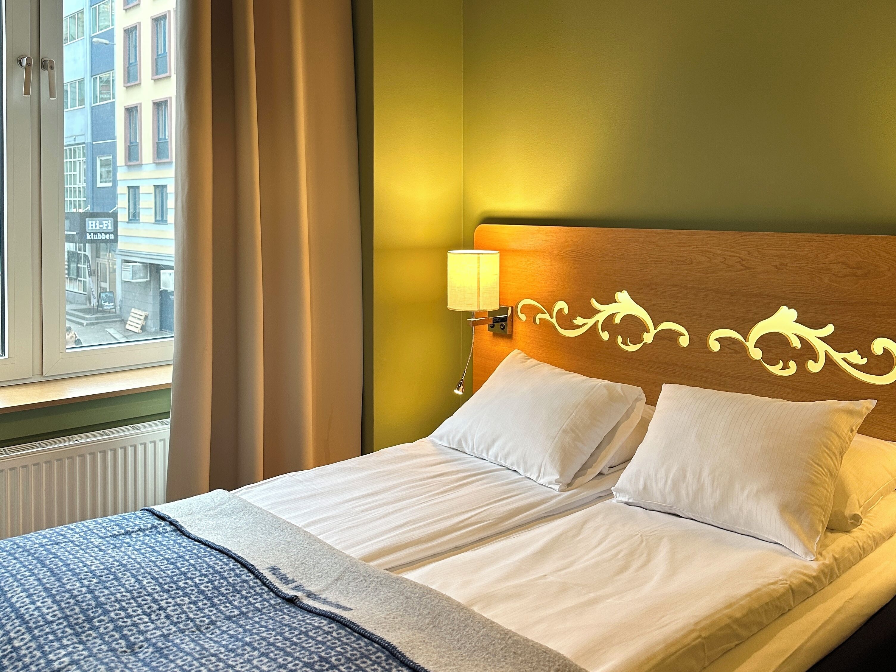 Habitación de hotel con cama, ventana con cortinas, lámpara de pared y vista a edificios.
