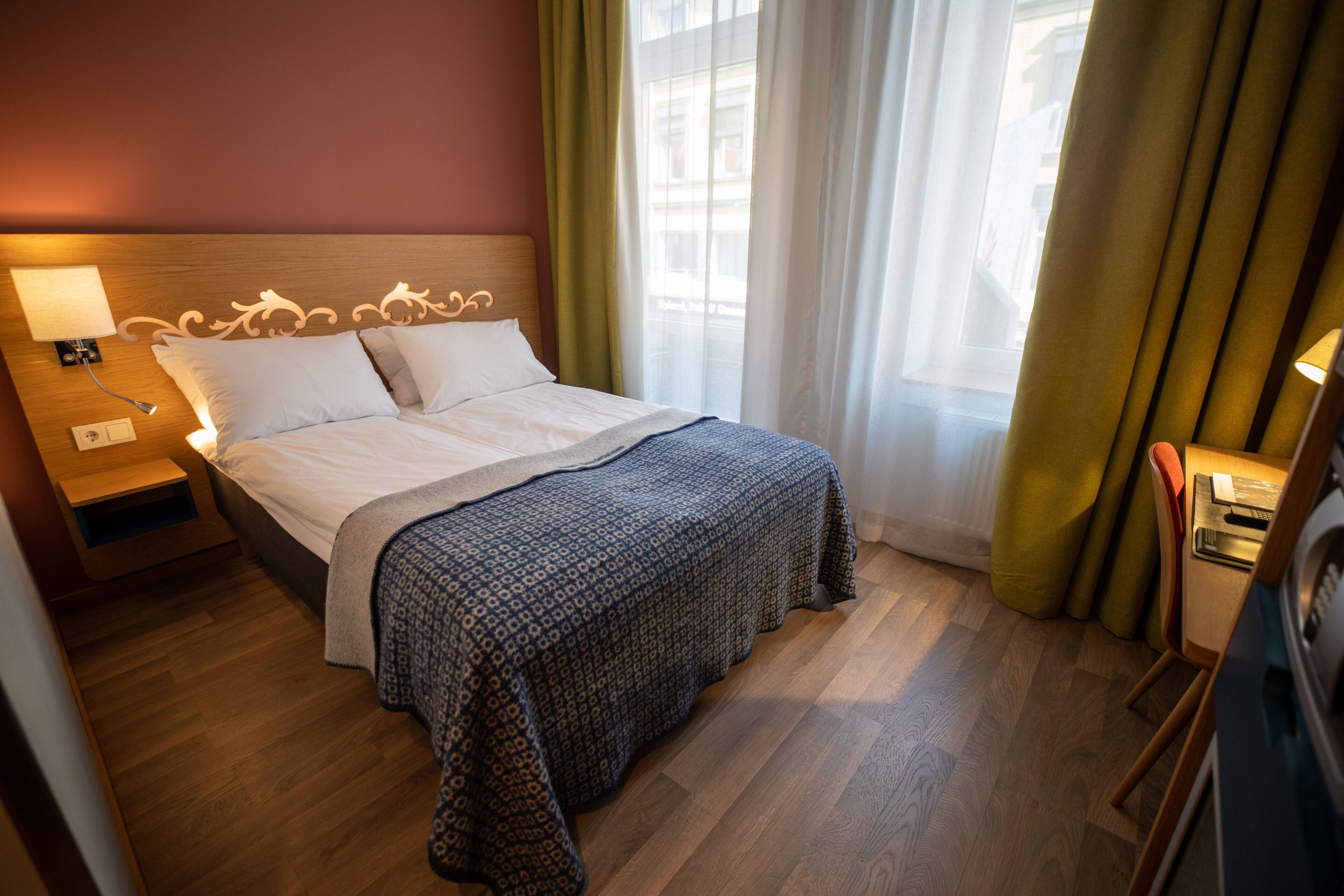 Una cama bien hecha con una colcha estampada, una mesita de noche con lámpara y vistas al exterior a través de una ventana con cortinas.