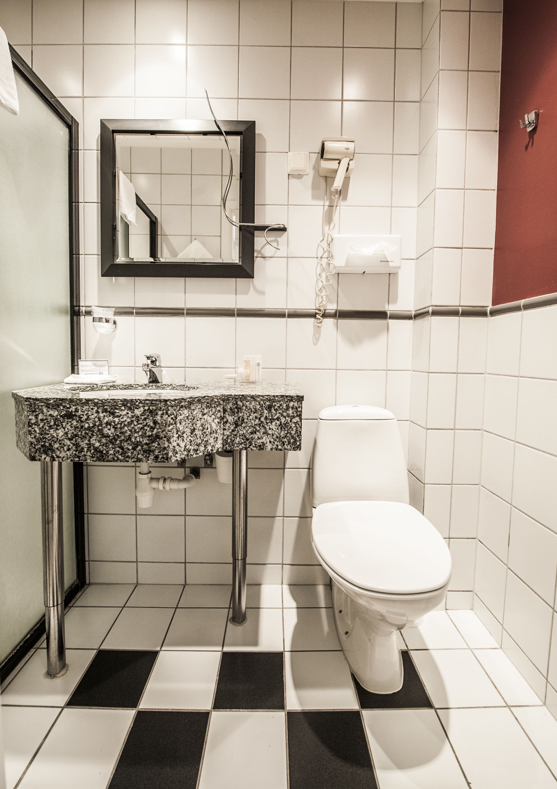 A bathroom with a white toilet, a sink with a granite countertop, a mirror, and a hairdryer mounted on the wall.