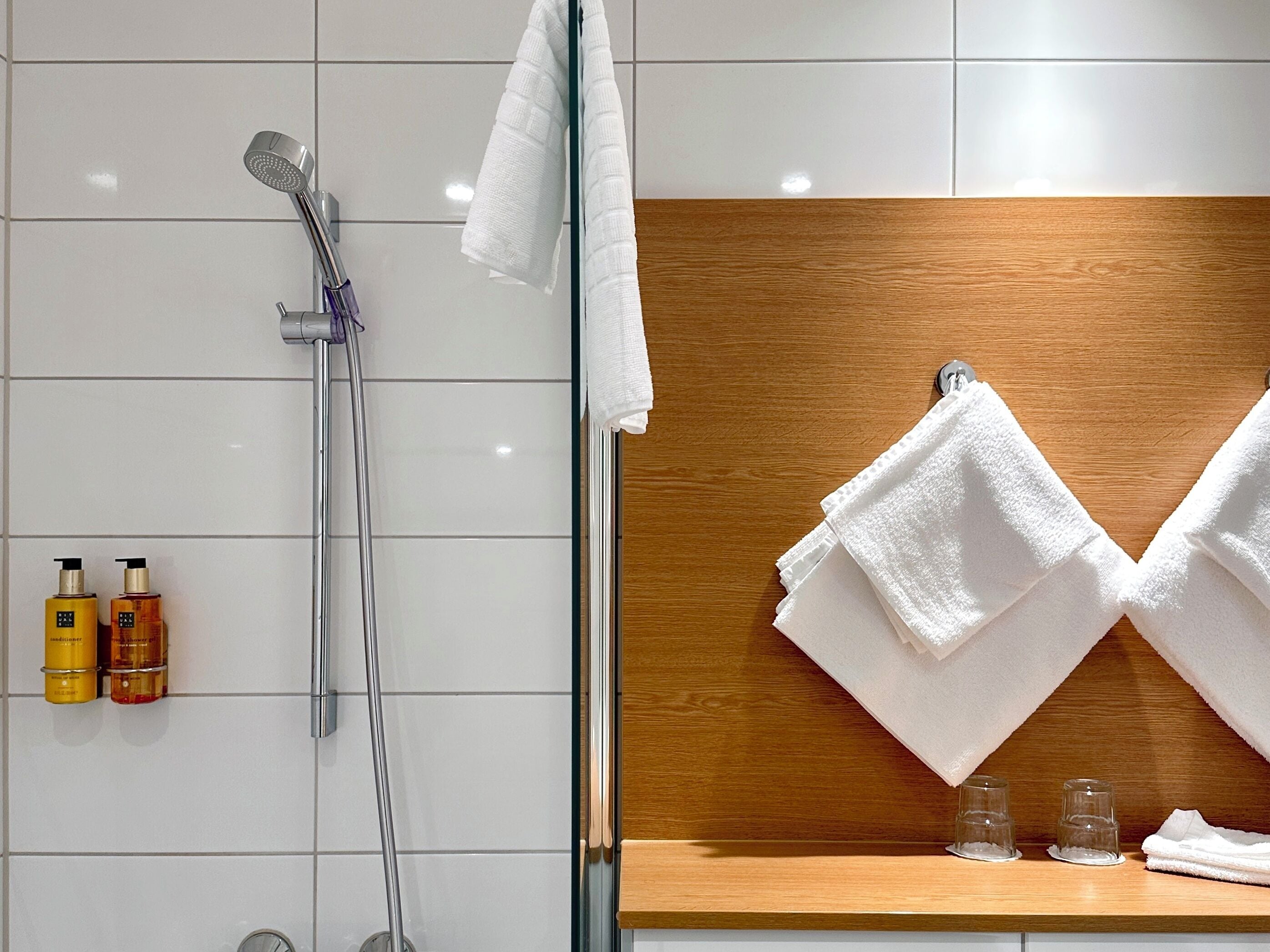 Een badkamer met witte tegels, een douche met een handdouche, twee flessen shampoo en twee handdoeken die aan een houten rek hangen.