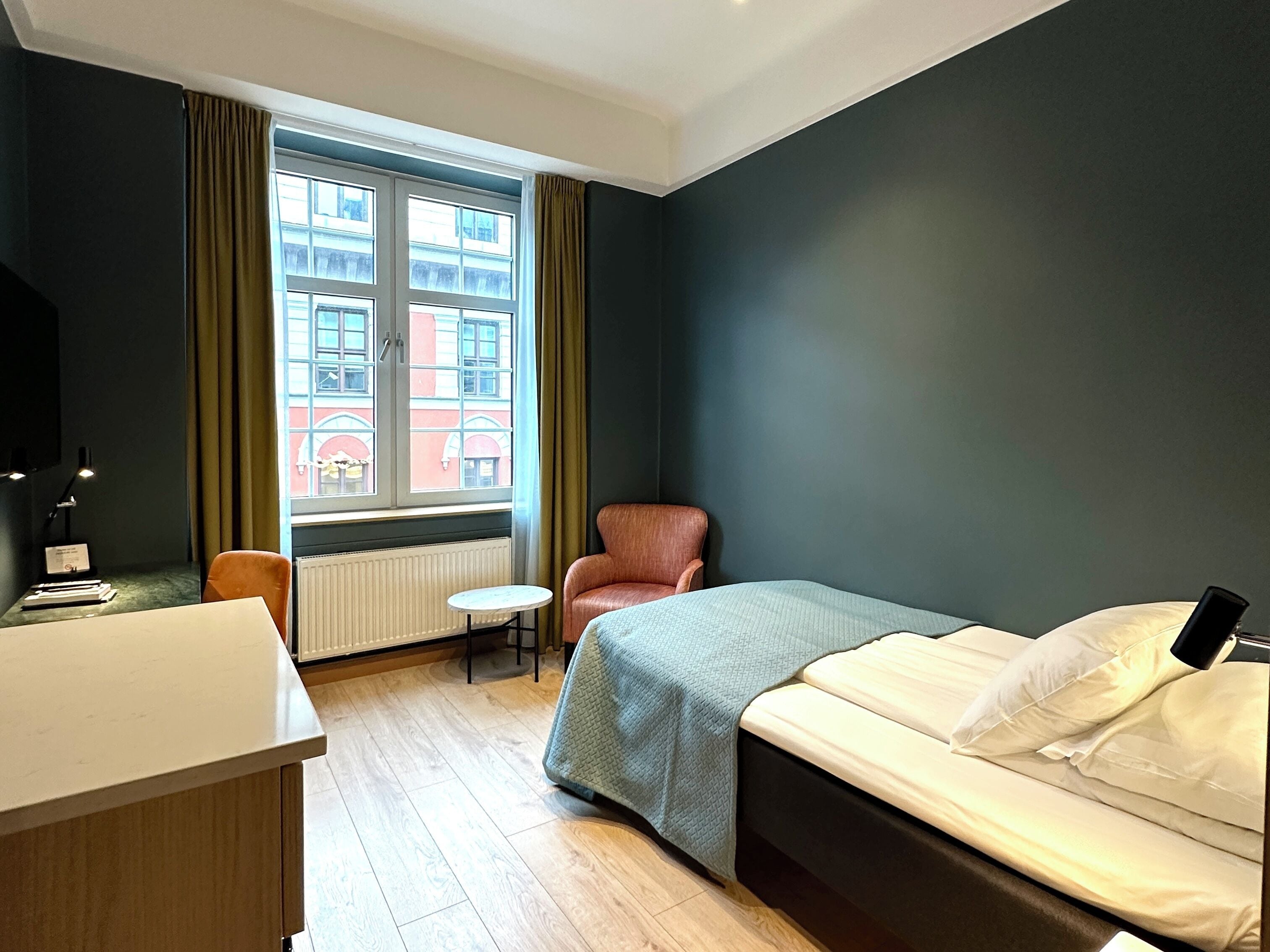 A hotel room with a bed, a desk, a chair, and a window with a view of buildings.