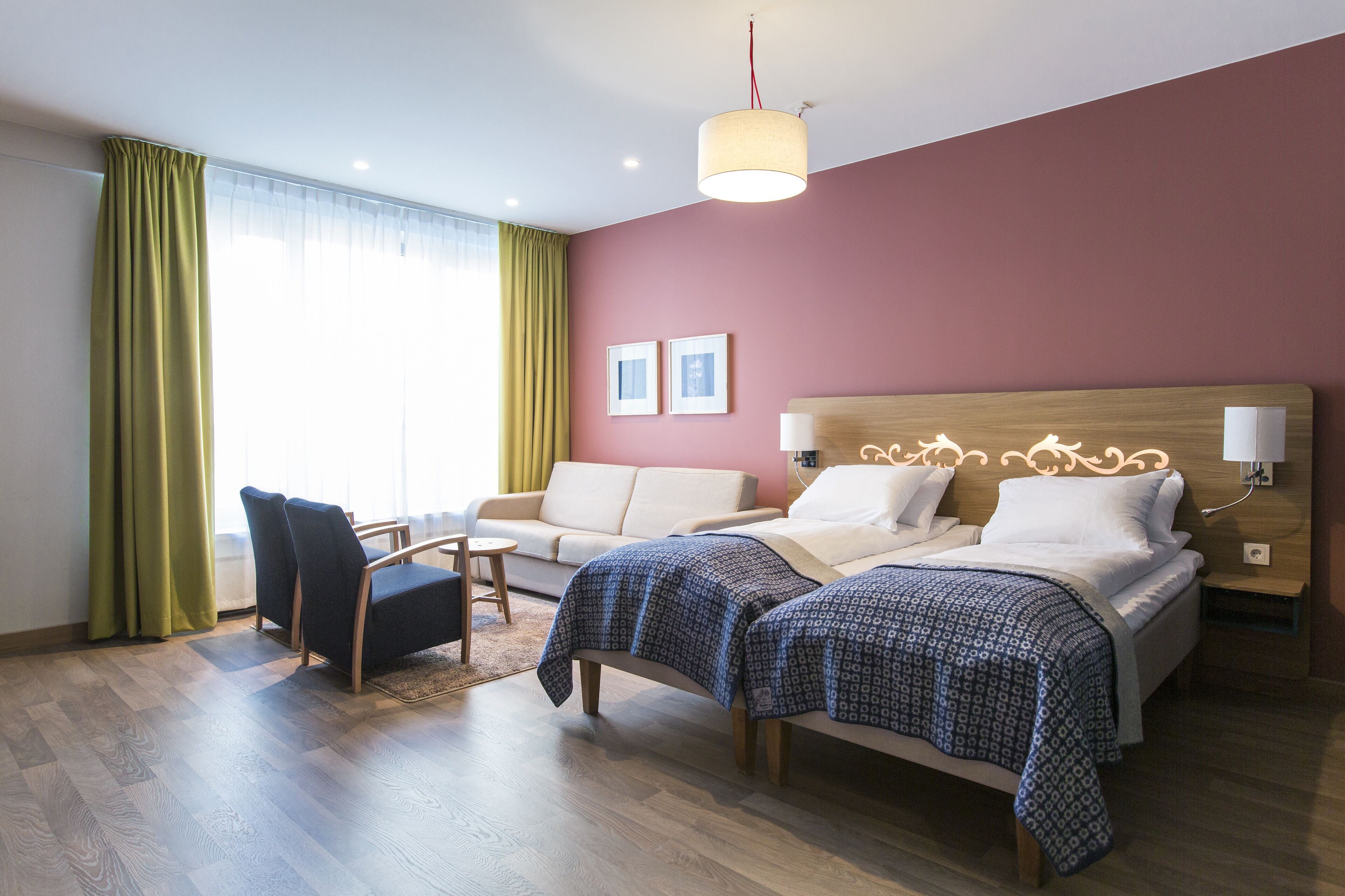 A hotel room with a bed, two bedside tables, a chair, a sofa, and a large window with curtains.