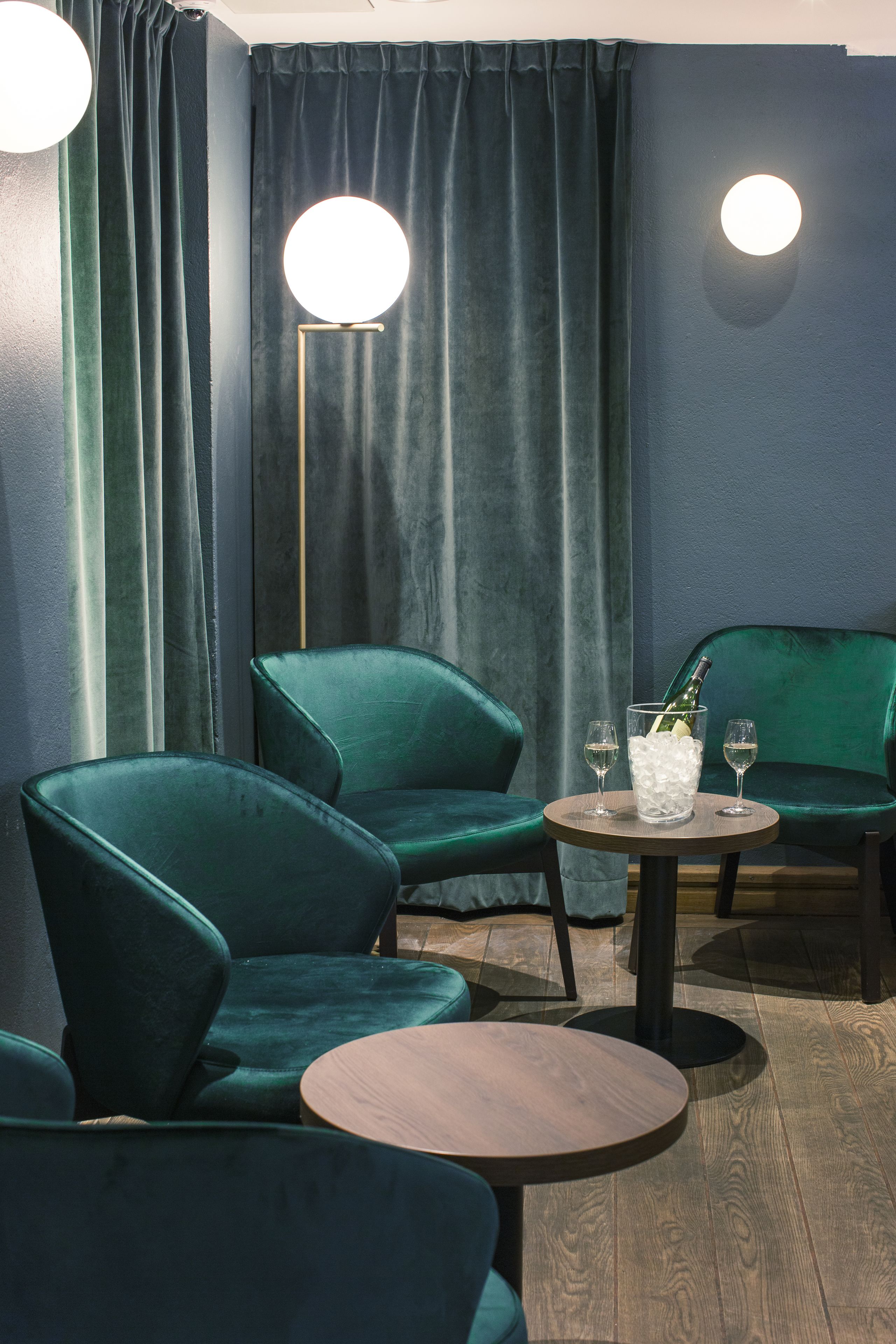A room with two green velvet armchairs, a round wooden table with a bottle and glasses, and a floor lamp.