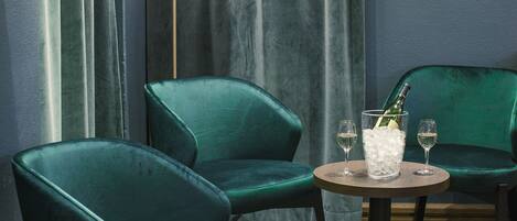 A room with two green velvet armchairs, a round wooden table with a bottle and glasses, and a floor lamp.