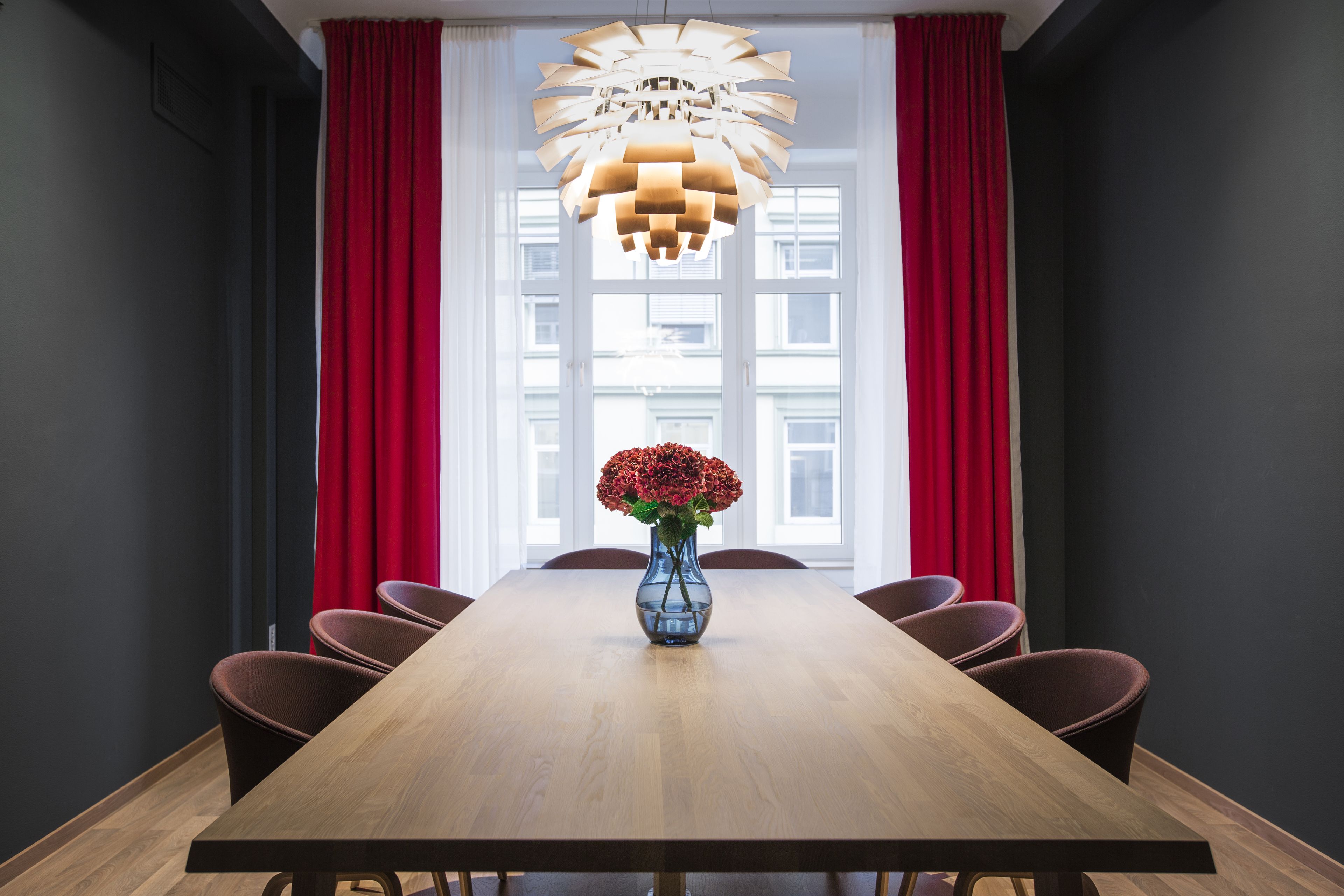Un salón de conferencias con una mesa de madera, cortinas rojas y un candelabro singular.